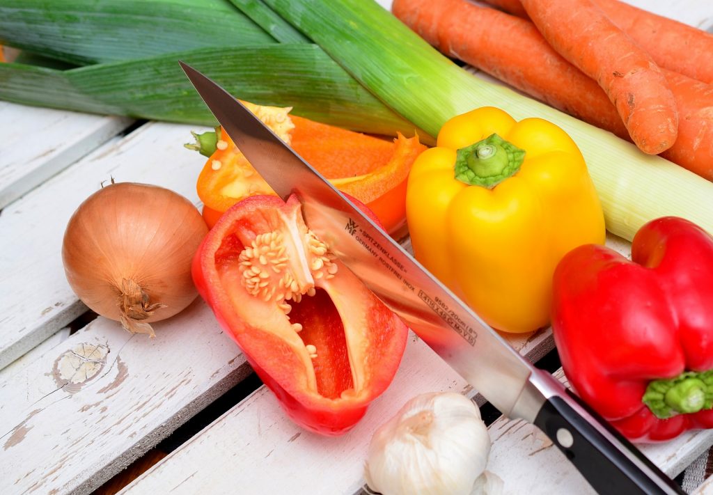 Knife cutting open a bell pepper, surrounded by a variety of vegetables on a wood table.