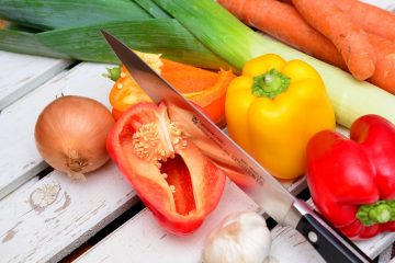 Knife cutting open a bell pepper, surrounded by a variety of vegetables on a wood table.