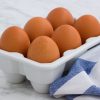 Carton of six brown eggs on white counter top with blue and white dishtowel.