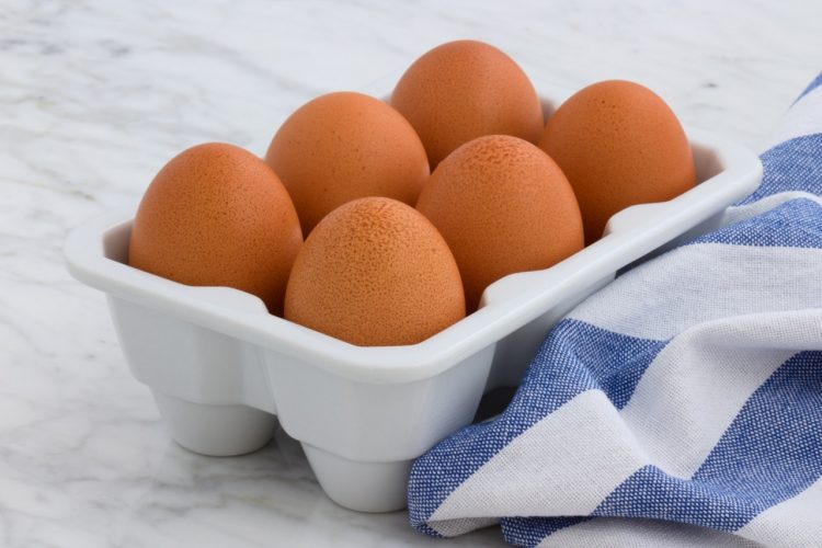 Carton of six brown eggs on white counter top with blue and white dishtowel.