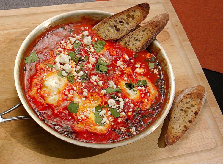 Shakshuka in a pan, paired with crispy bread and topped with feta cheese and cilantro.