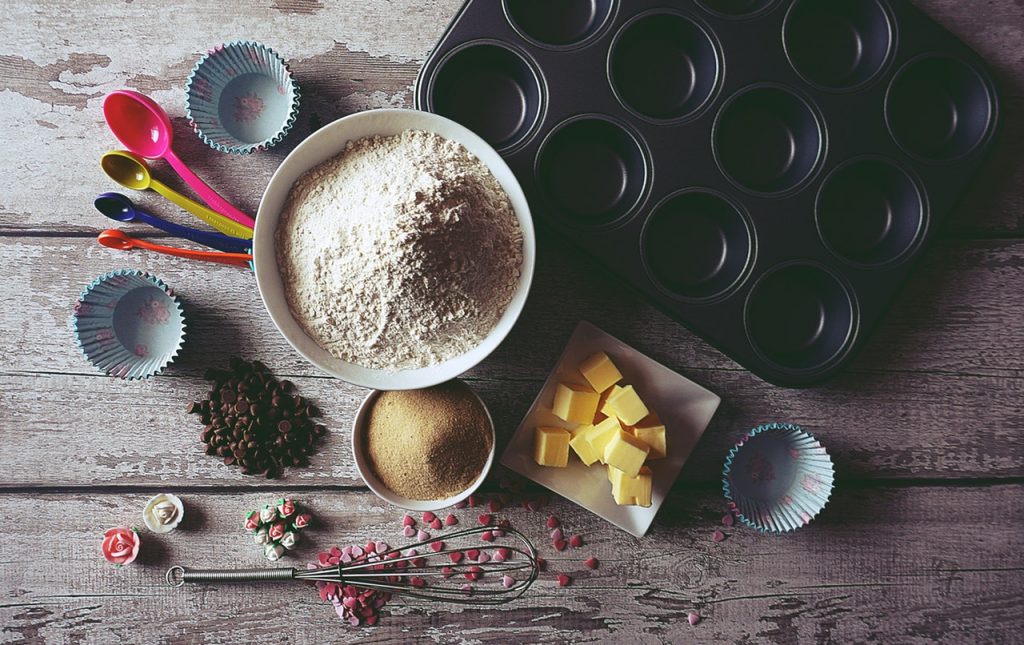 Variety of baking items on a wooden table, including flour, sugar, butter, whisk, muffin tins, muffin cups and measuring spoons.