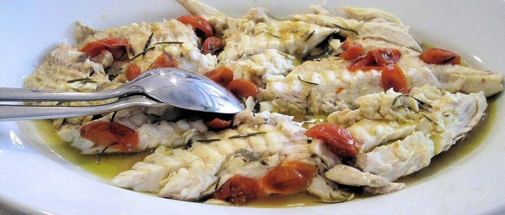 Cooked white fish in a tomato olive oil sauce on a plate with spoon beside it.