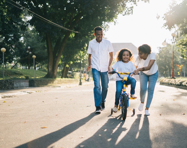 Father and mother helping younger sister ride bike with training wheels in neighborhood street.